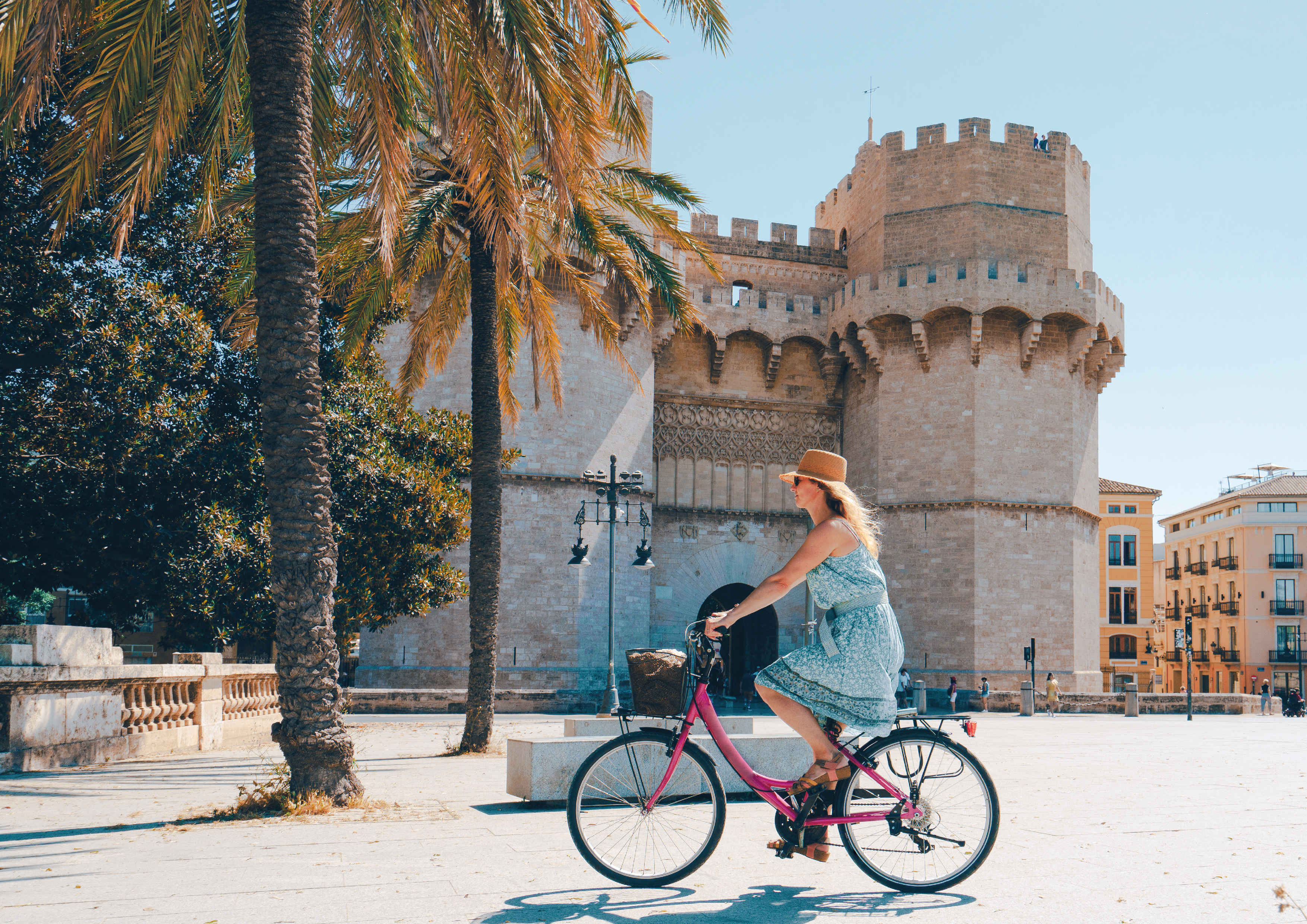 Frau auf einem pinken Fahrrad und mit Sonnenhut fährt entspannt durch eine Altstadt